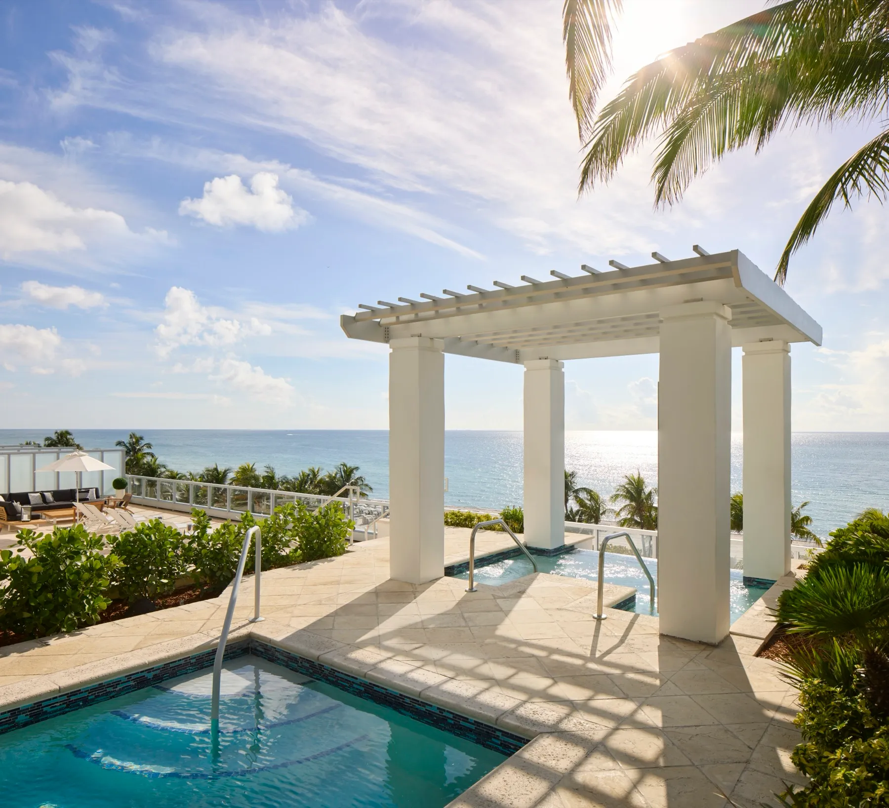 Sunny rooftop terrace with a white pergola over a hot tub, a small plunge pool, palm trees and a wide ocean view